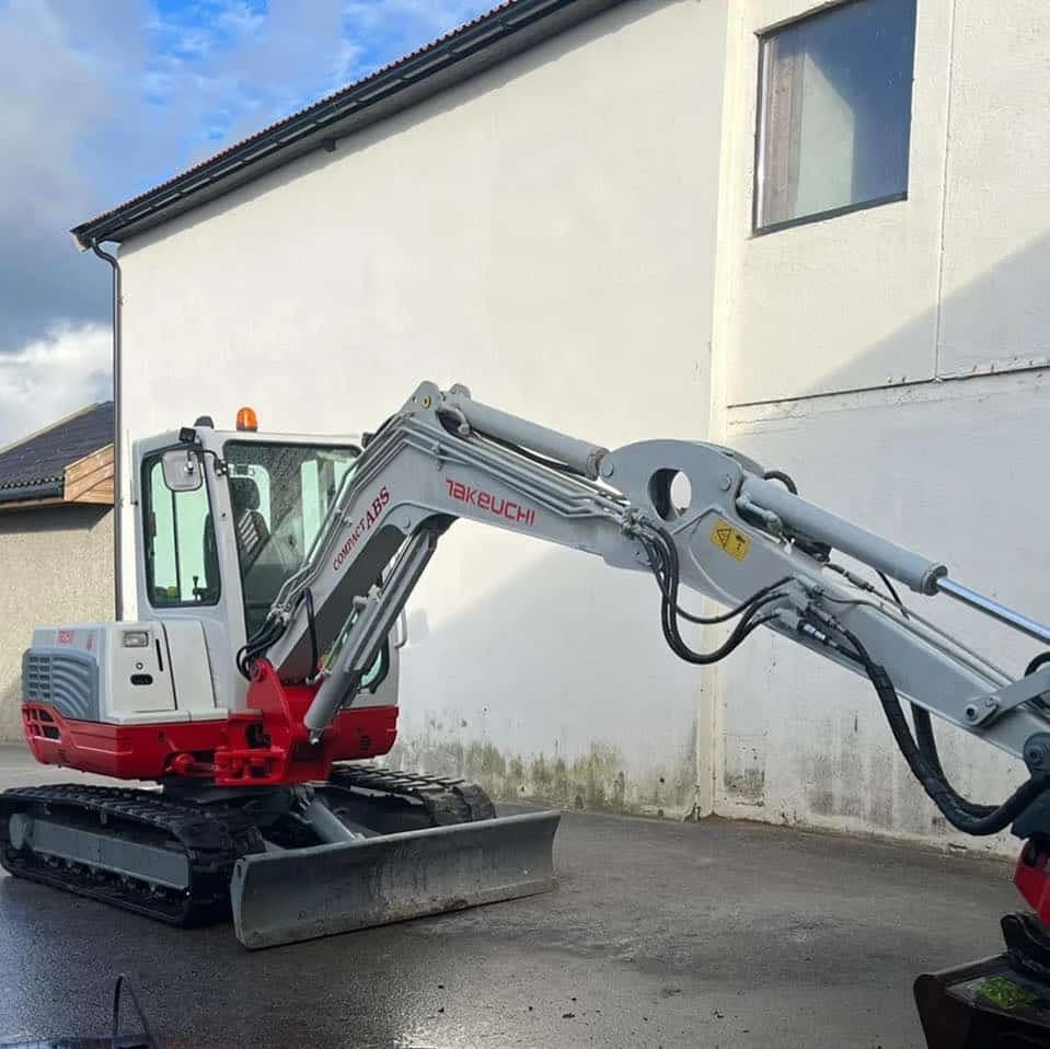 Kompakt gravemaskin parkert på asfalt ved en husvegg.