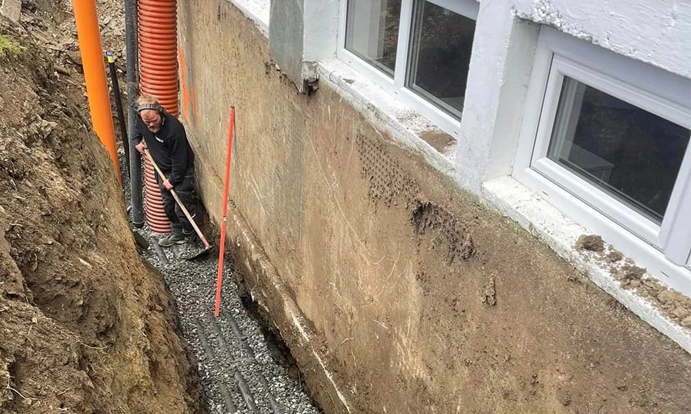 Person arbeider med rake i pukk i en grøft ved avdekket grunnmur og kjellervinduer.